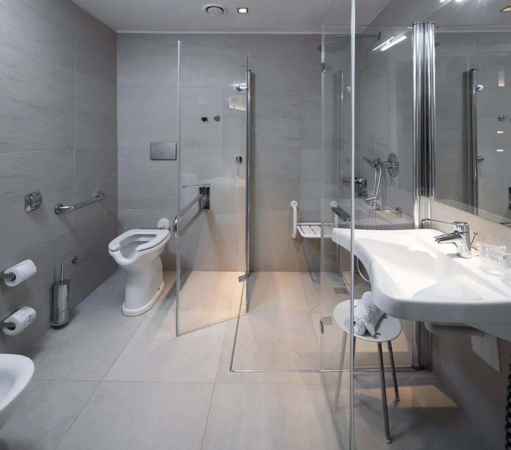 The image shows the accessible hotel room bathroom. On the right is the white suspended sink and behind it is the large shower cubicle with doors that open almost completely. Inside the shower cubicle is a folding stool on the wall. Next to the cubicle is the toilet.