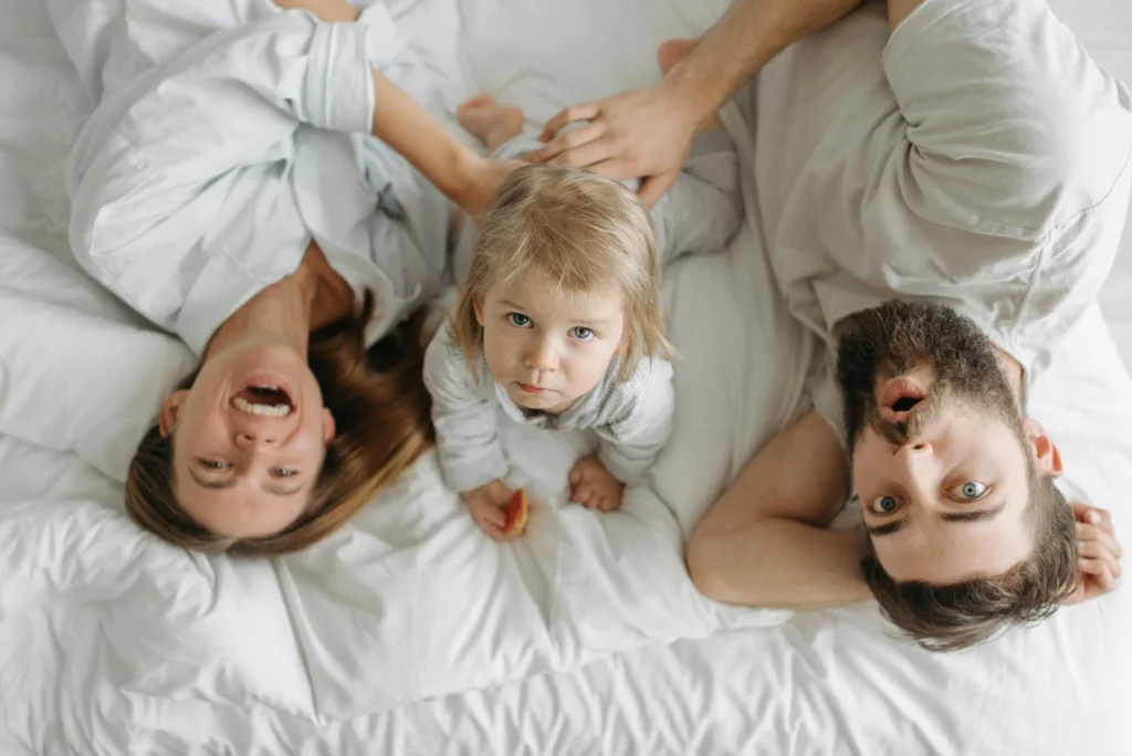 Nell'immagine una donna, un uomo e una bambina sono sdraiati su un letto. La donna sorride, l'uomo fa una faccia stupita e la bimba guarda in alto.