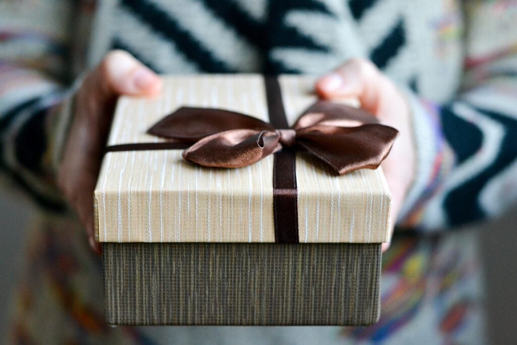 Nell'immagine due mani tengono un pacco regalo. Il pacco è marrone scuro e il coperchio è beige con un grande fiocco marrone.